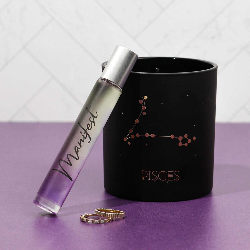 Manifest Rollerball Perfume bottle with purple ombre design next to a black Pisces constellation candle and gold rings on a purple surface
